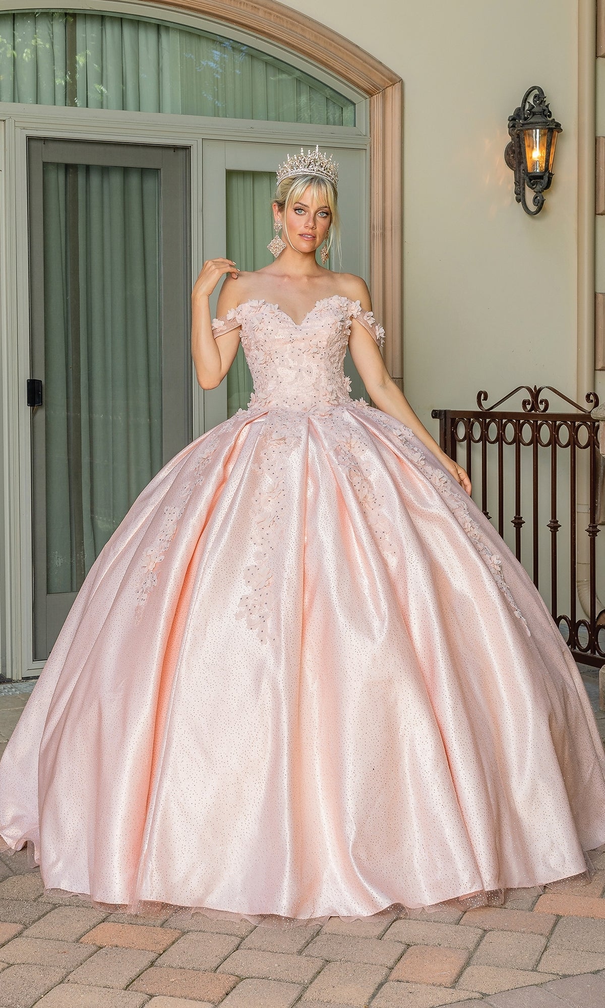 Someone stands on a stone patio before glass doors and a wrought iron railing, wearing the Quinceanera Dress 1501 by Dancing Queen—a pink off-the-shoulder ball gown with a sweetheart neckline and a decorative crown.